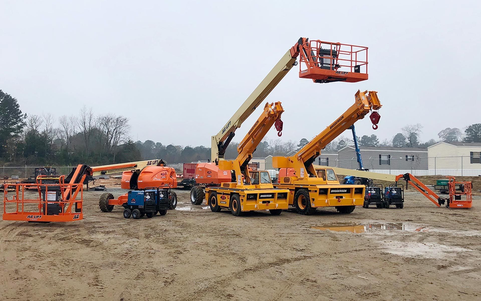 Construction site with several orange and yellow boom lifts on muddy ground.