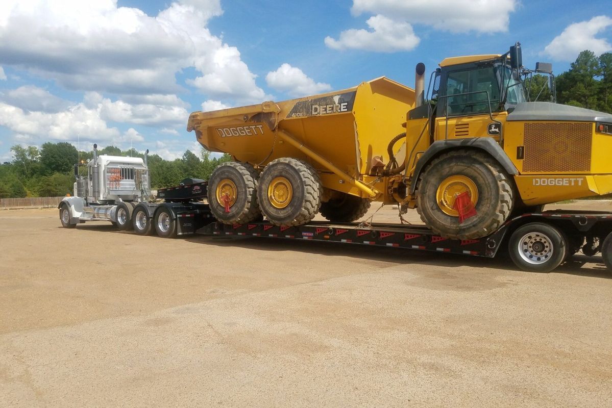 Semi-truck hauling a large yellow articulated dump truck on a flatbed trailer on a gravel lot under a blue sky.