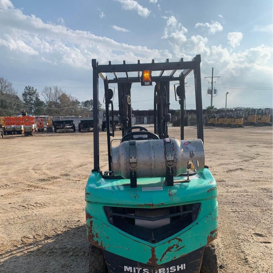 Green Mitsubishi forklift outdoors, propane tank visible, with a warning light on, on a dirt lot.