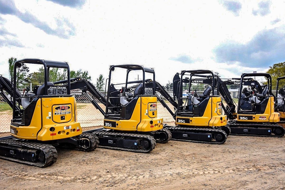 Four yellow mini-excavators parked in a row on a dirt surface under a cloudy sky.