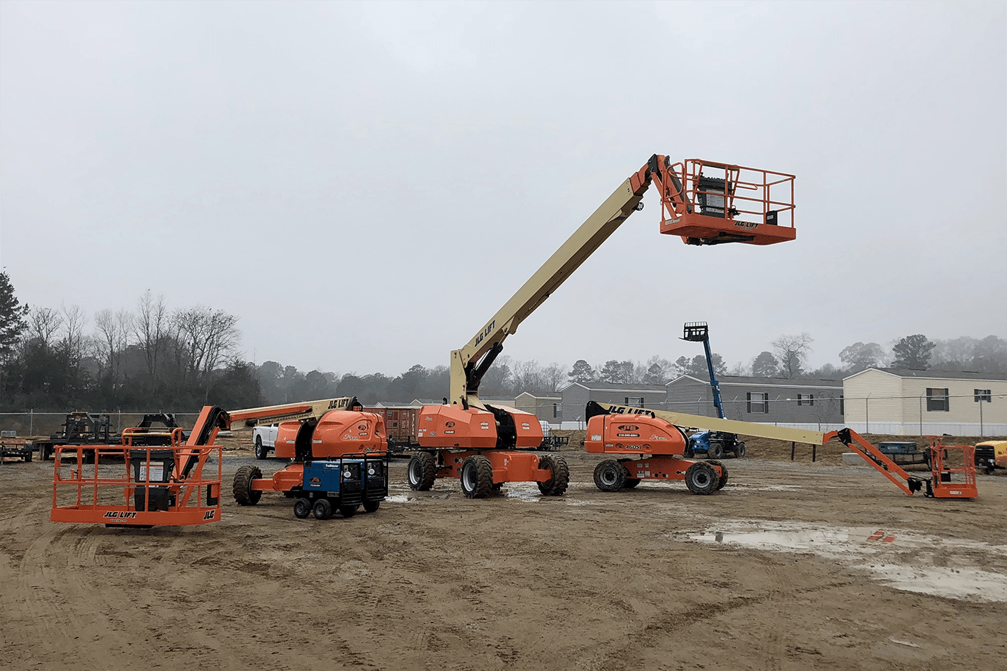 Several orange boom lifts on a muddy construction site under overcast skies.