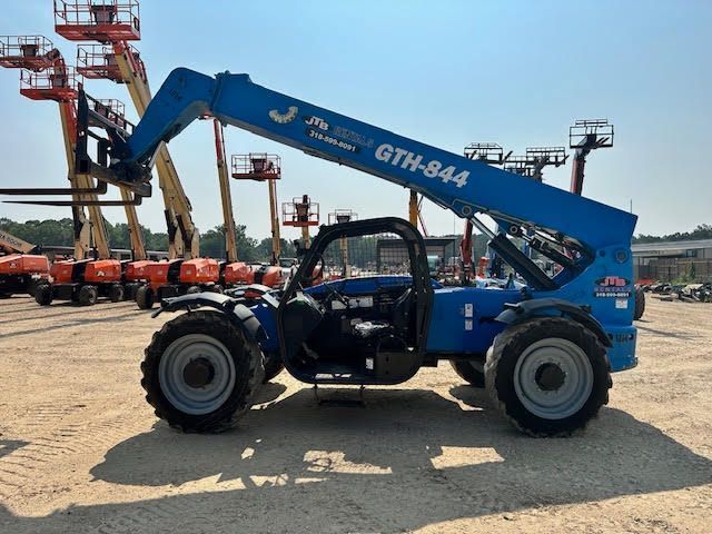 Blue Genie GTH-844 telehandler with extended boom on dirt, other machinery in background under a sunny sky.