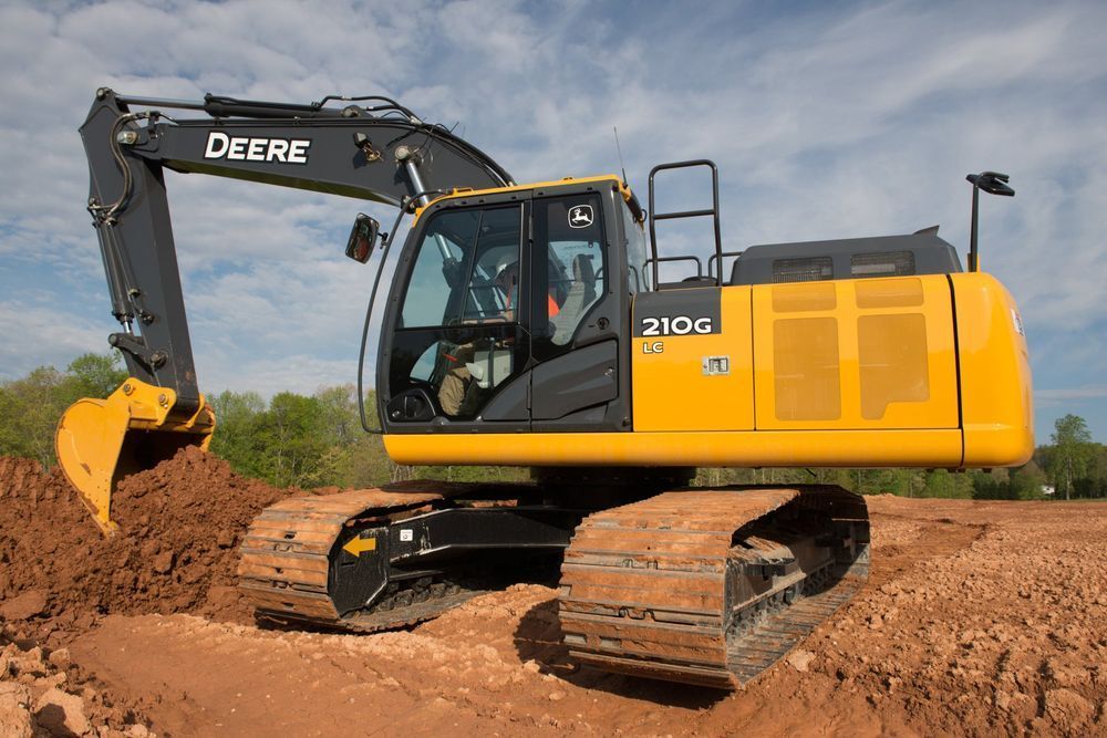 John Deere 210G excavator digging in a dirt field. Yellow and black machine.