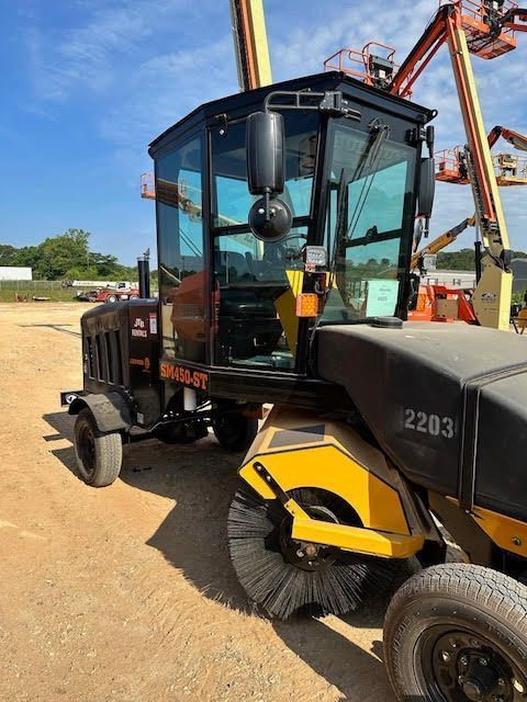 Street sweeper vehicle with a cab and rotating brushes outdoors.