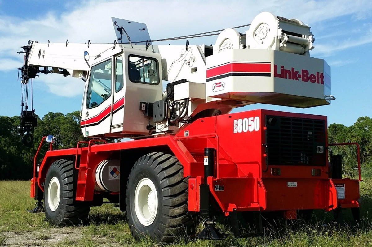 Red and white Link-Belt crane, parked on grass. The crane has large wheels, a cab, and a boom.