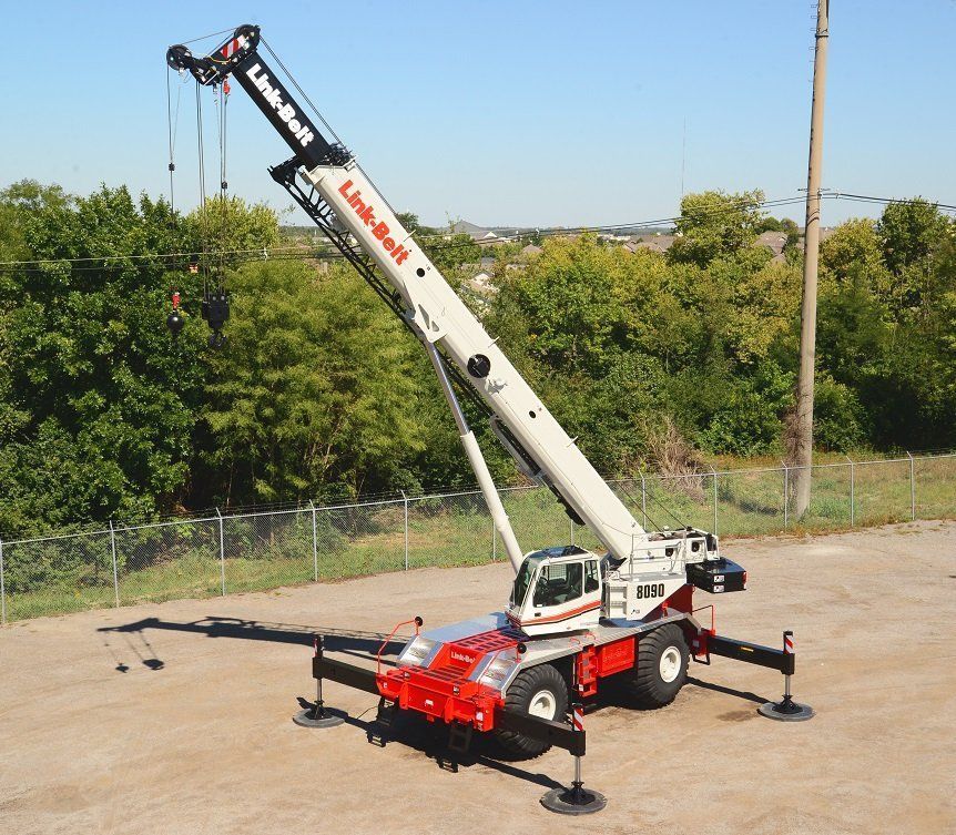 A white and red Link-Belt crane on a construction site with trees and sky in the background.