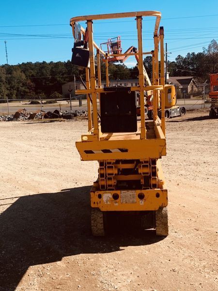 Yellow scissor lift on a gravel surface under a blue sky, with trees and buildings in the background.