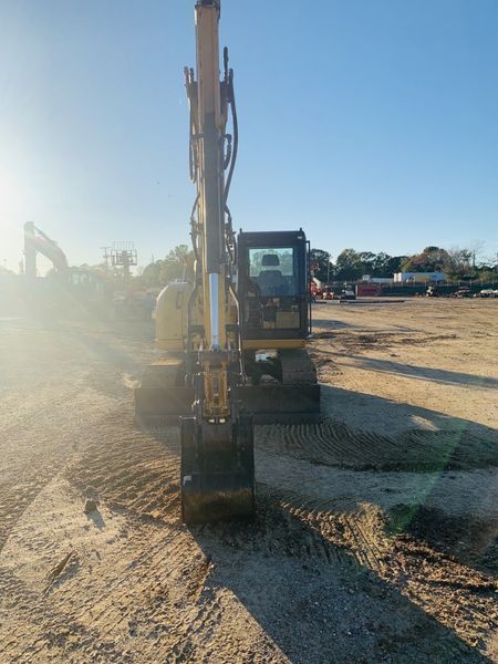 Yellow excavator on a dirt lot, sunlight, cab with operator, bucket facing forward.
