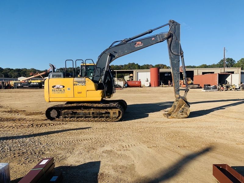 Yellow and black excavator on a dirt lot with metal structures in the background.