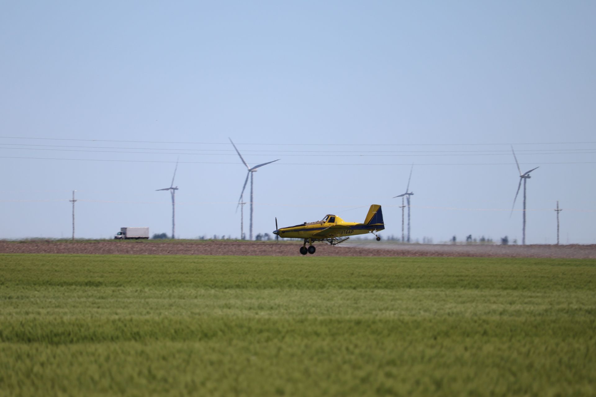 A yellow and black plane is taking off from a runway in a field with wind turbines in the background.