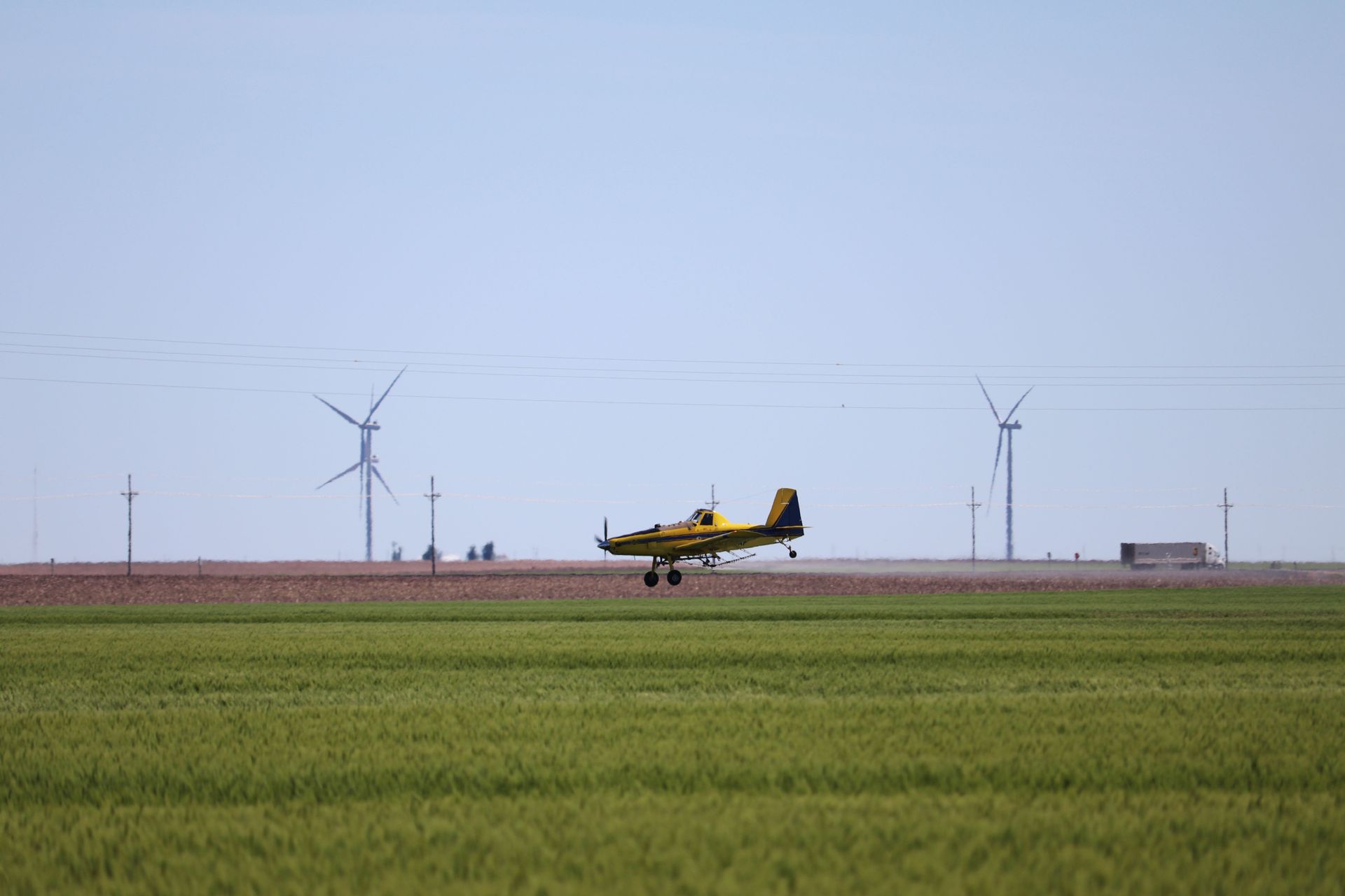 A yellow plane is taking off from a grassy field with windmills in the background.