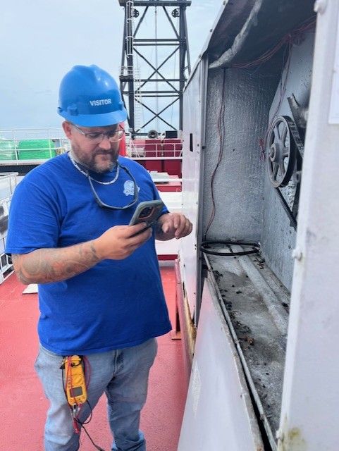 HVAC technician in blue hard hat and shirt inspecting unit on a rooftop, using a phone.
