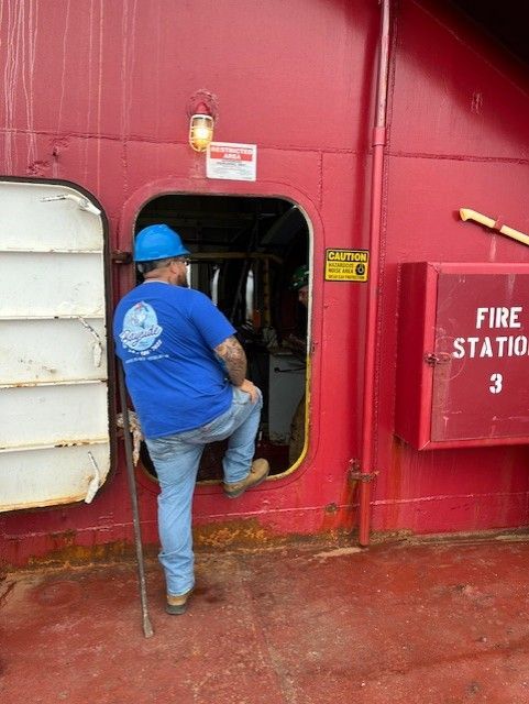 Man in blue hard hat and shirt steps into a doorway on a red ship, holding a cane.