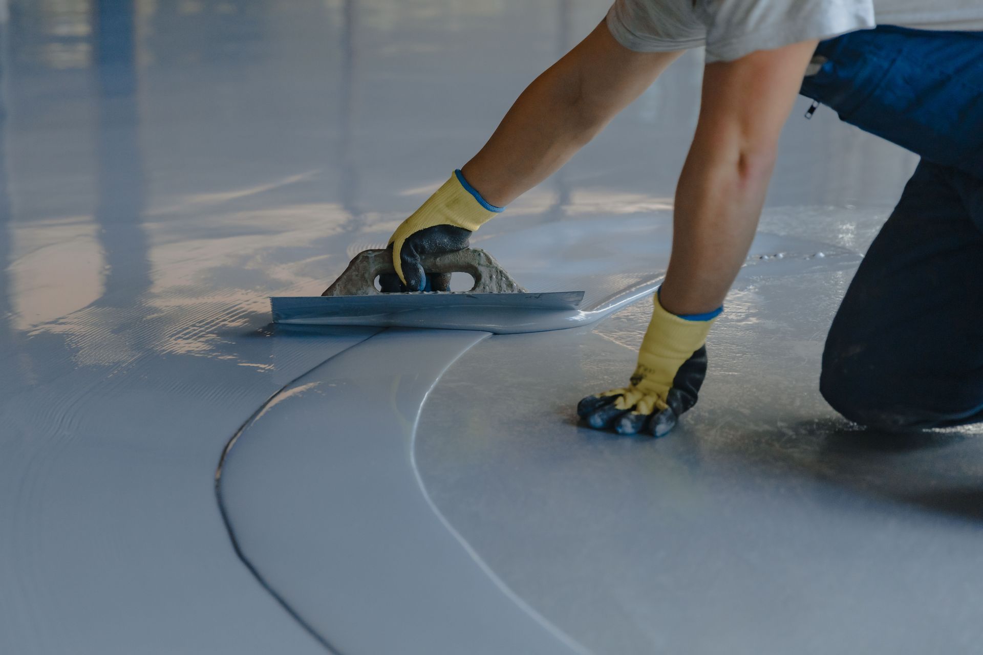 Worker smoothing a gray floor coating with a trowel, wearing gloves and kneeling on the surface