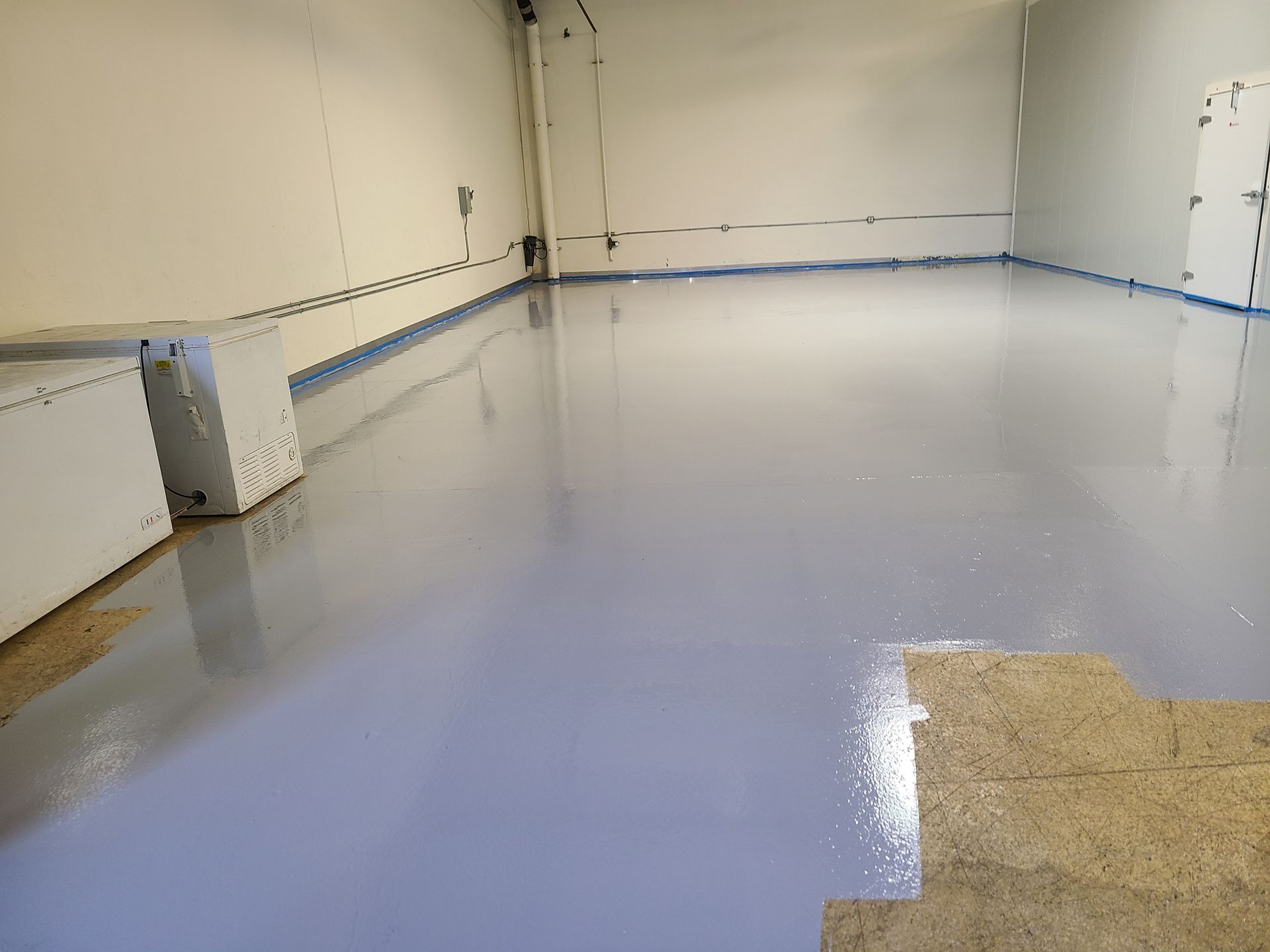 Empty room with a glossy light-blue floor and beige walls, partially covered by cardboard in the corner