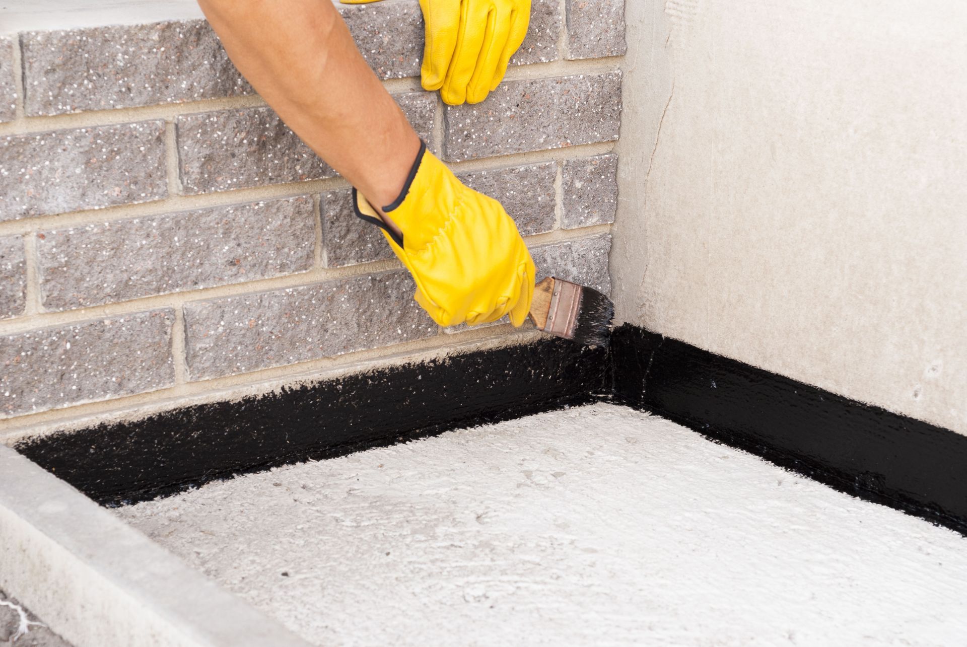Gloved hands applying black sealant along the edge where a brick wall meets a floor corner