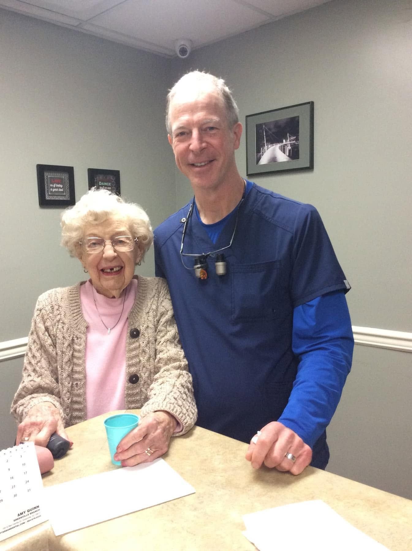 Two smiling people stand at a table in a gray room, one seated and one standing beside her.