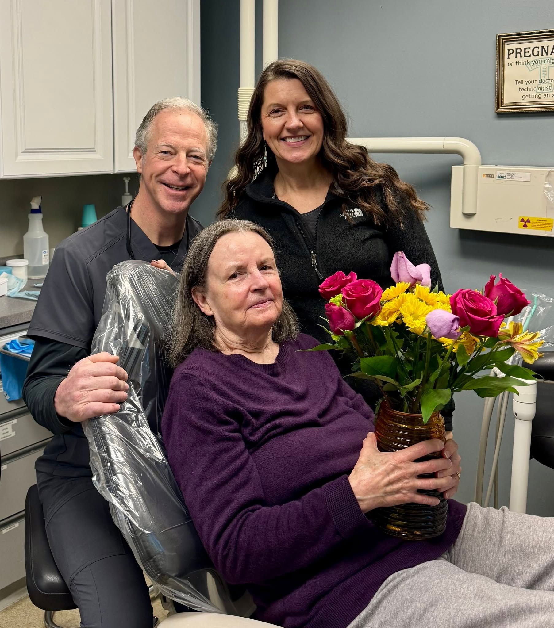 Three people in a dental office, smiling, with one woman holding a bouquet of bright flowers.