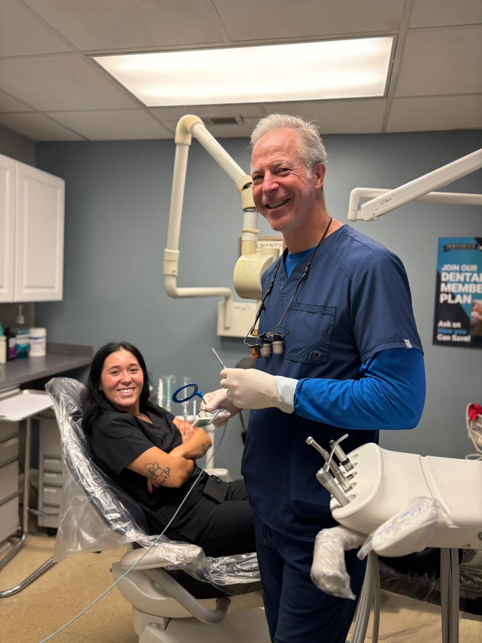 Dental patient smiling in chair while dentist in blue scrubs stands beside her in a clinic room.