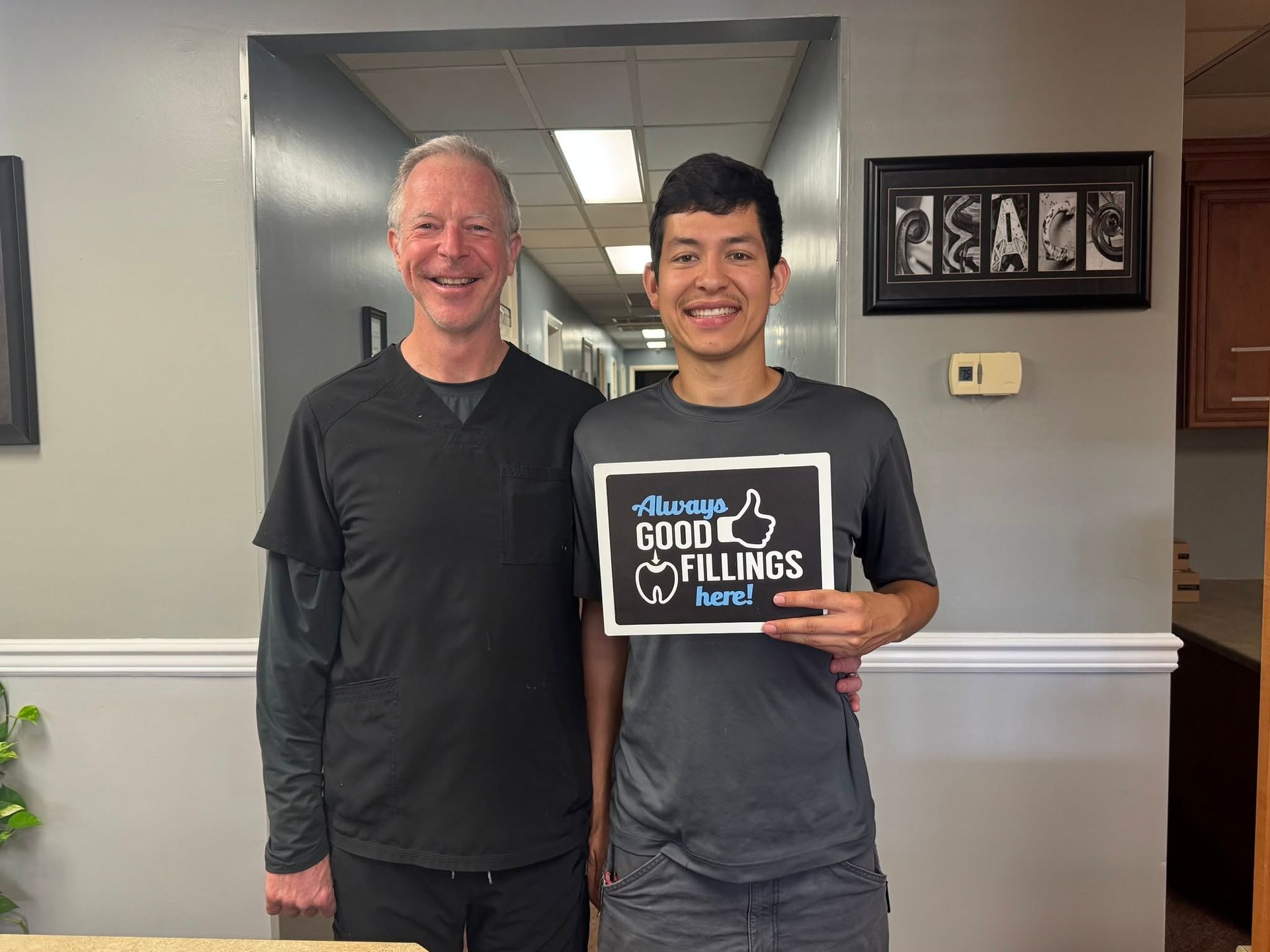 Two people smiling indoors, one holding a plaque, standing in a room with a window and wall art.