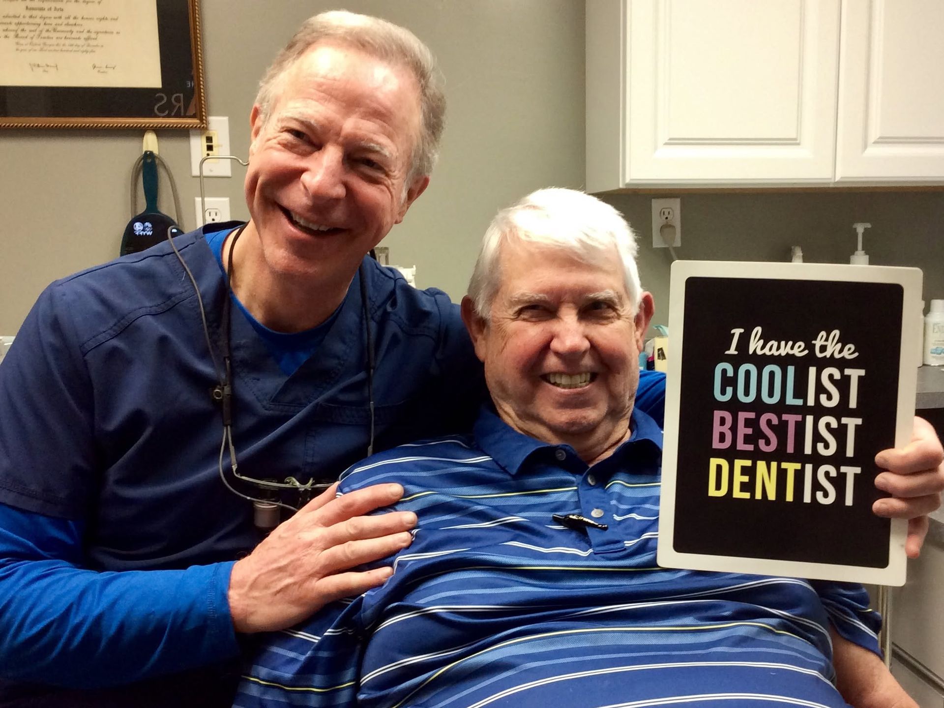 Two smiling men in blue shirts pose in a dental office, holding a sign that says “I have the coolest bestest dentist.”