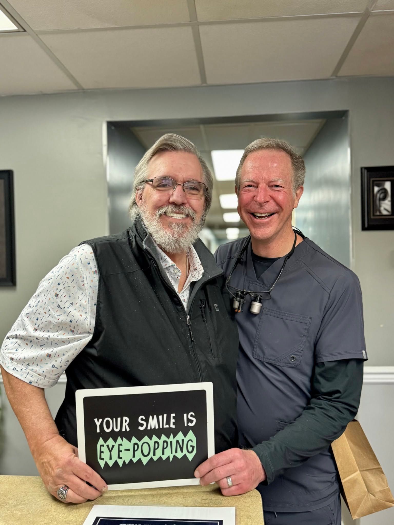 Two smiling men indoors hold a sign that says “Your smile is yippy popping.”