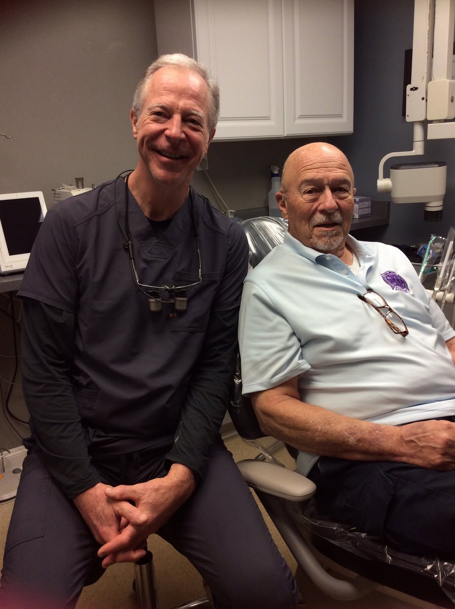 Two smiling people seated in a medical office, one in a chair and one on a rolling stool, with cabinets behind them