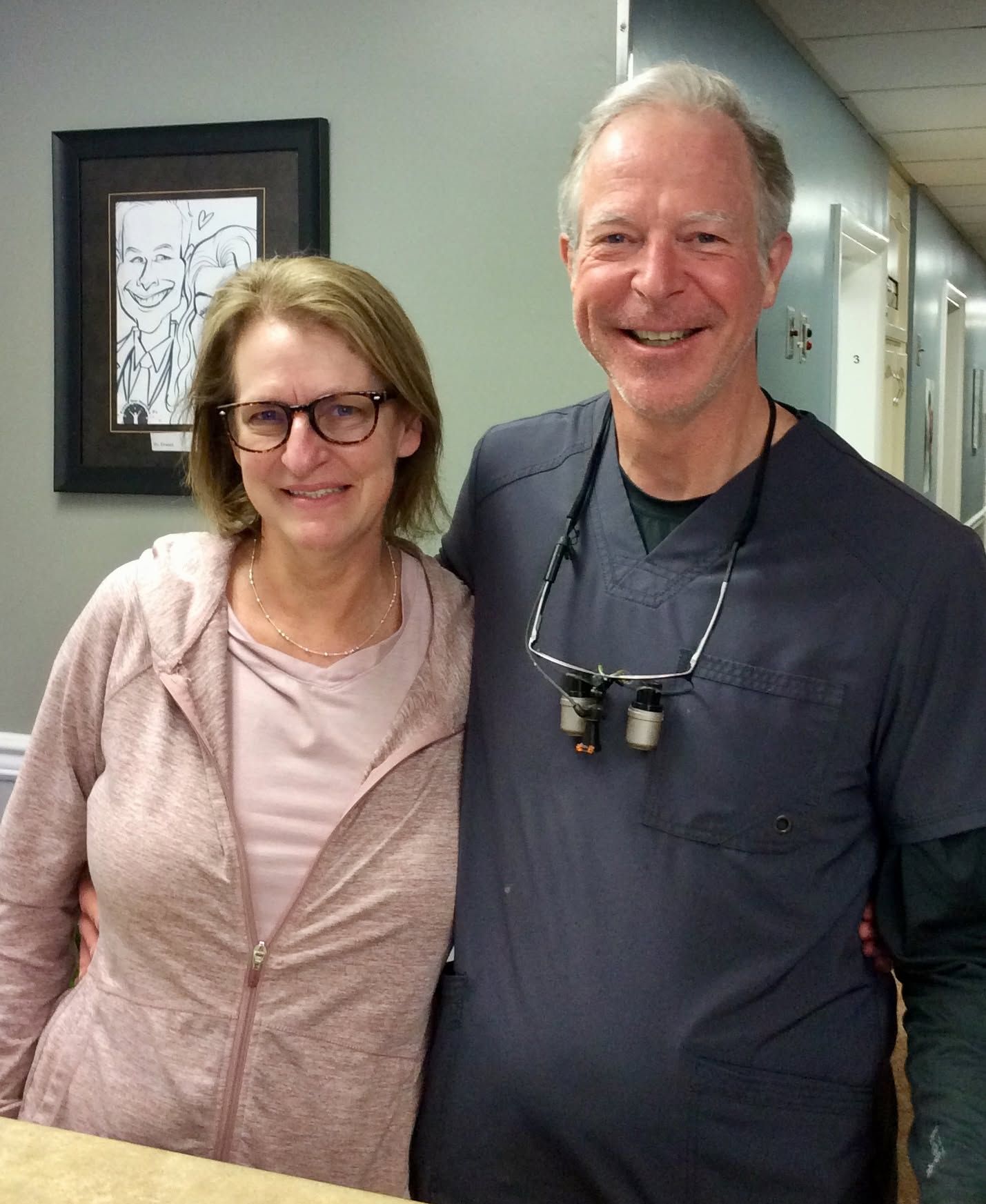 Two smiling people standing in a hallway, with framed wall art behind them.