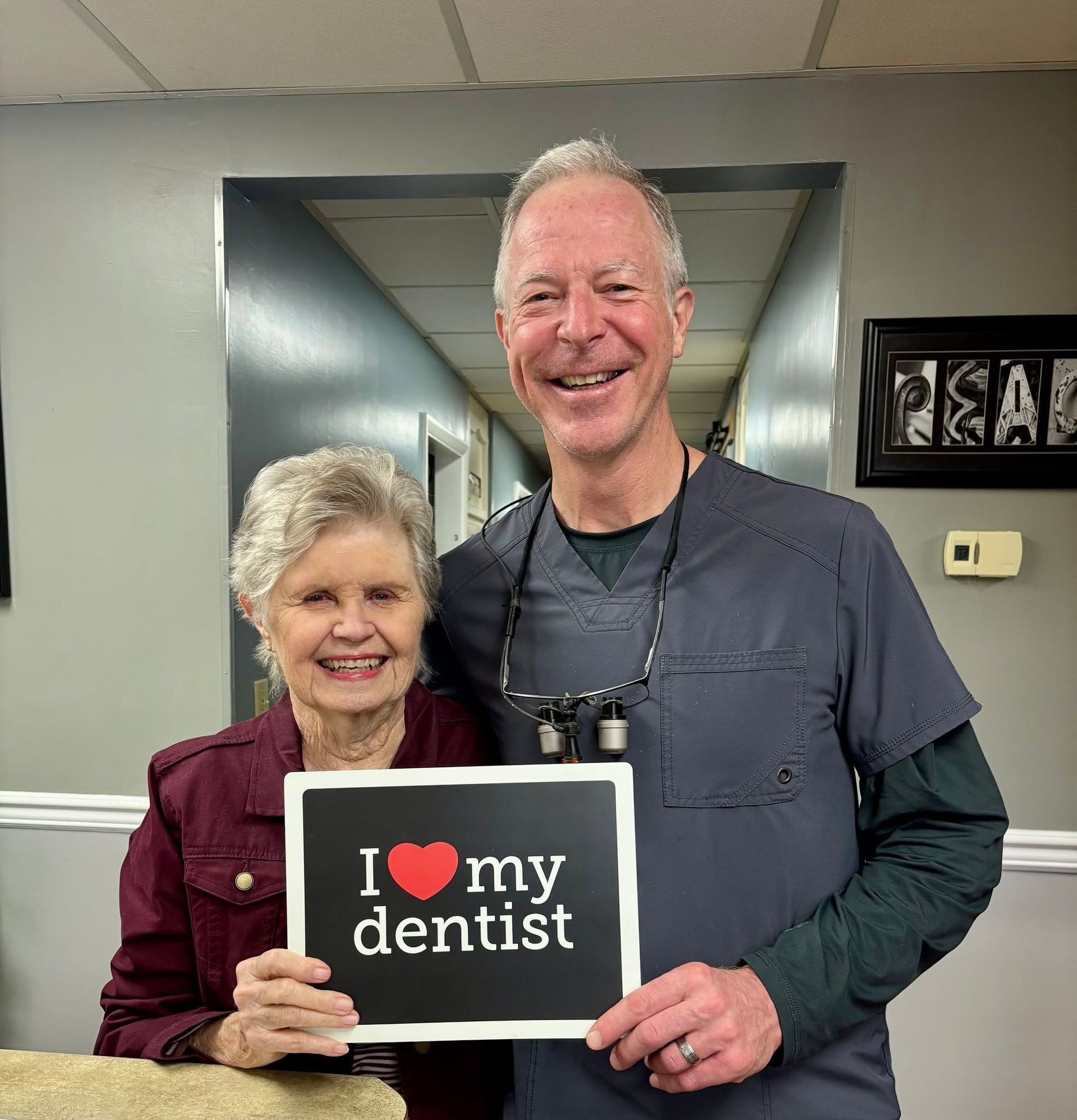 Two people smiling and holding a sign that says “I ♥ my dentist” in a dental office hallway