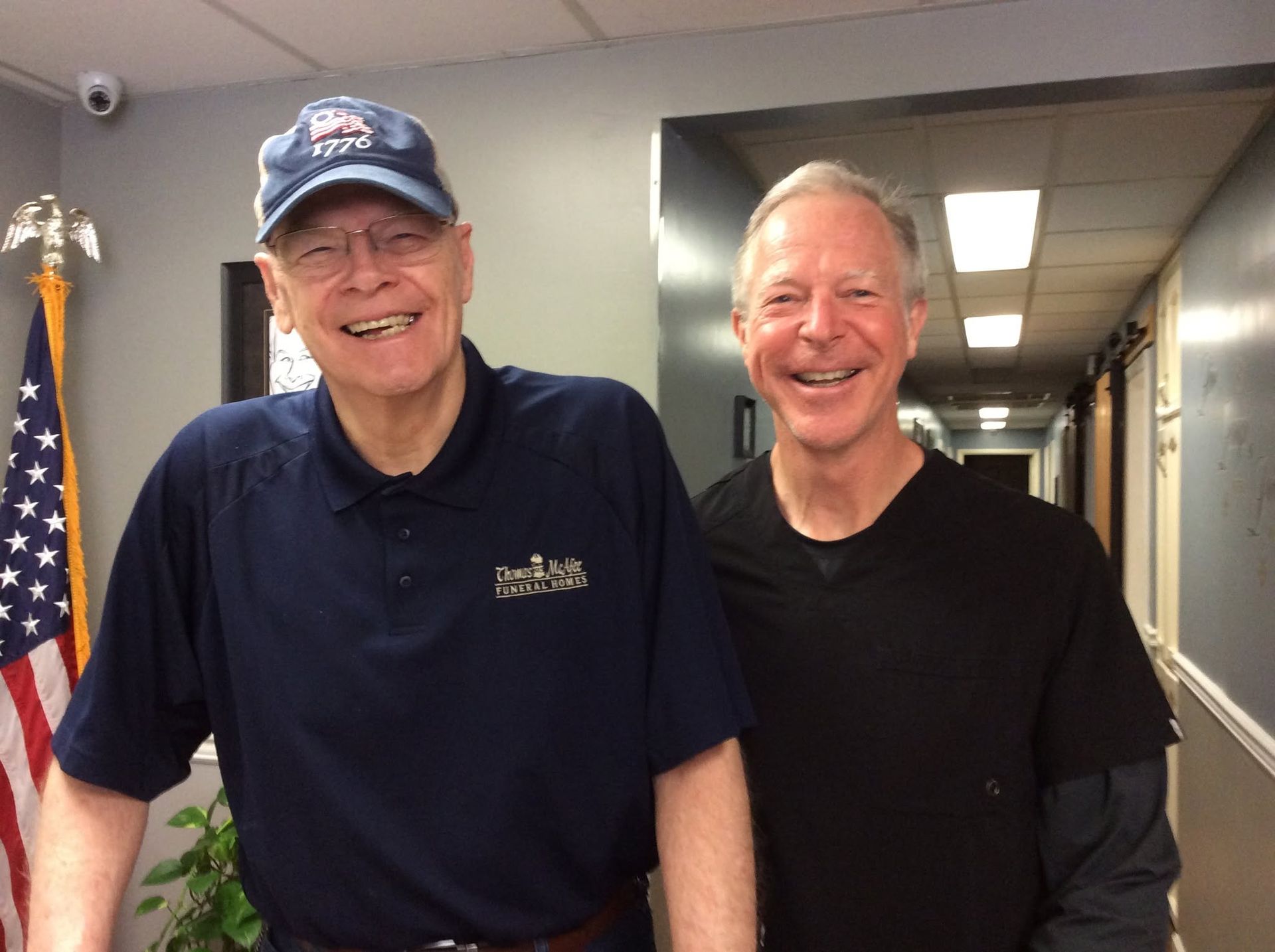 Two smiling men stand in a hallway, one in a navy cap and shirt, the other in a black shirt.