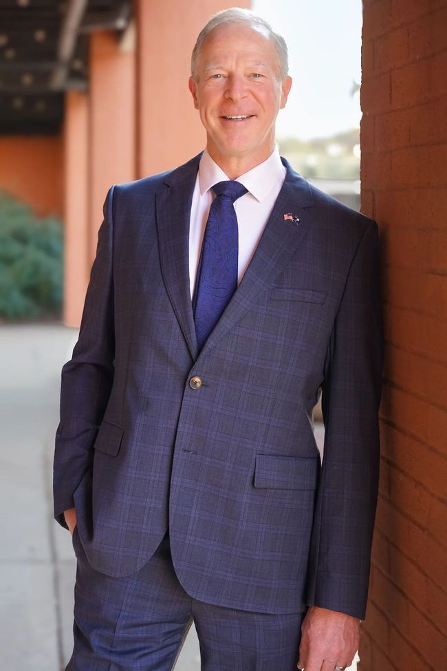 Man in a navy suit standing outdoors beside a brick wall, smiling at the camera.