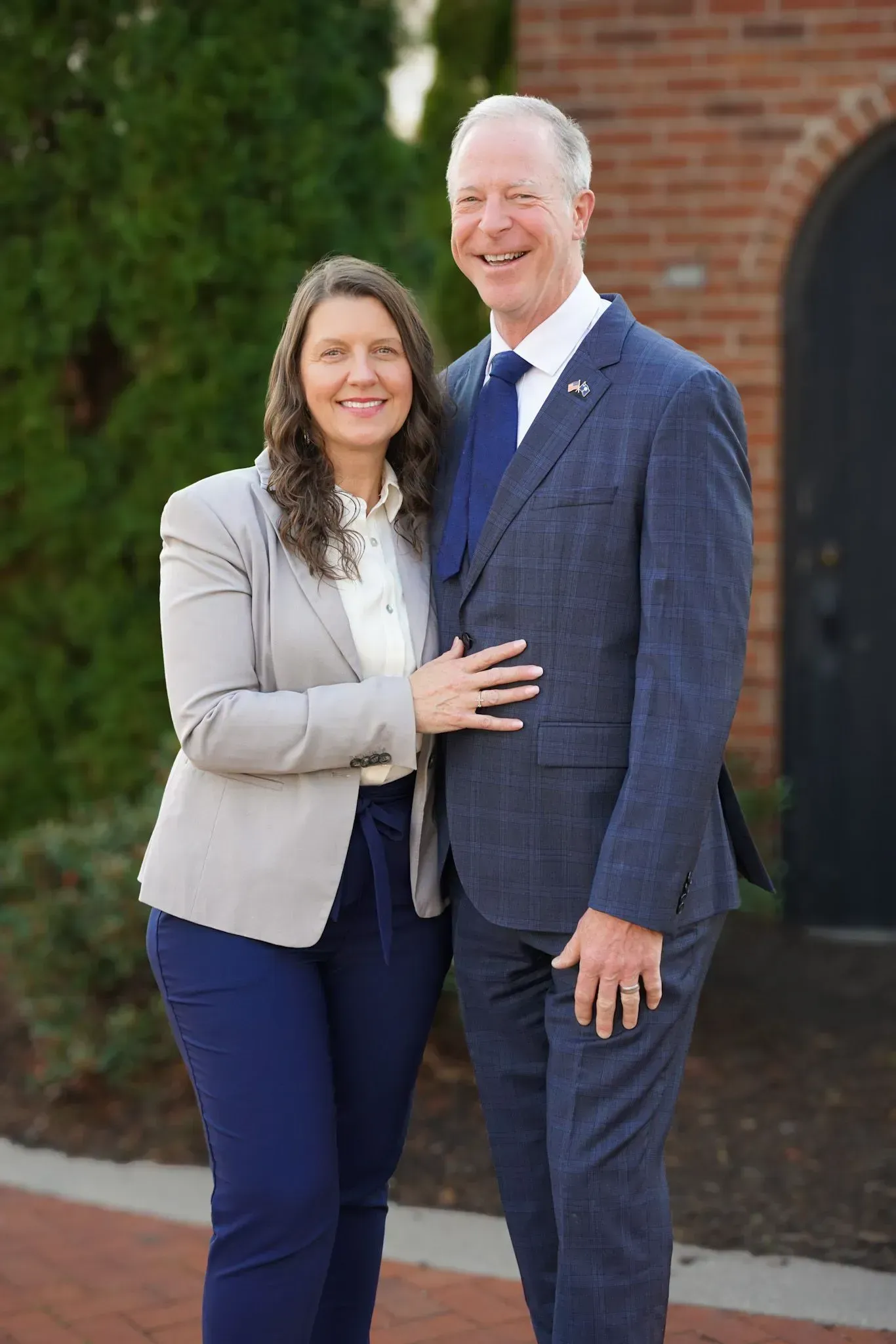 Two people smiling and standing together outdoors by a brick building, dressed in business attire