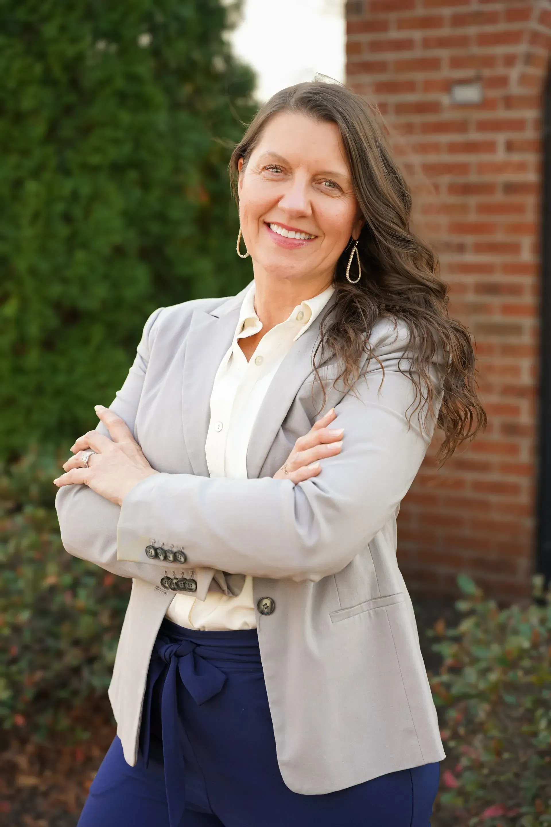 Woman standing outdoors with arms crossed, wearing a light blazer and dark pants, smiling near a brick wall.