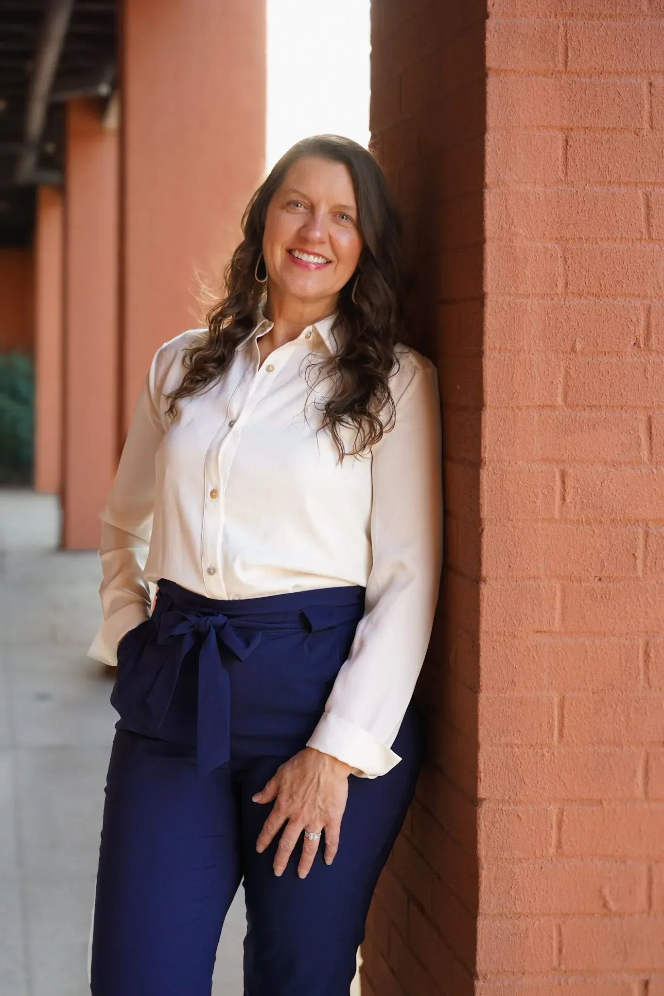 Woman in white blouse and navy pants leaning against a brick wall outdoors, smiling.