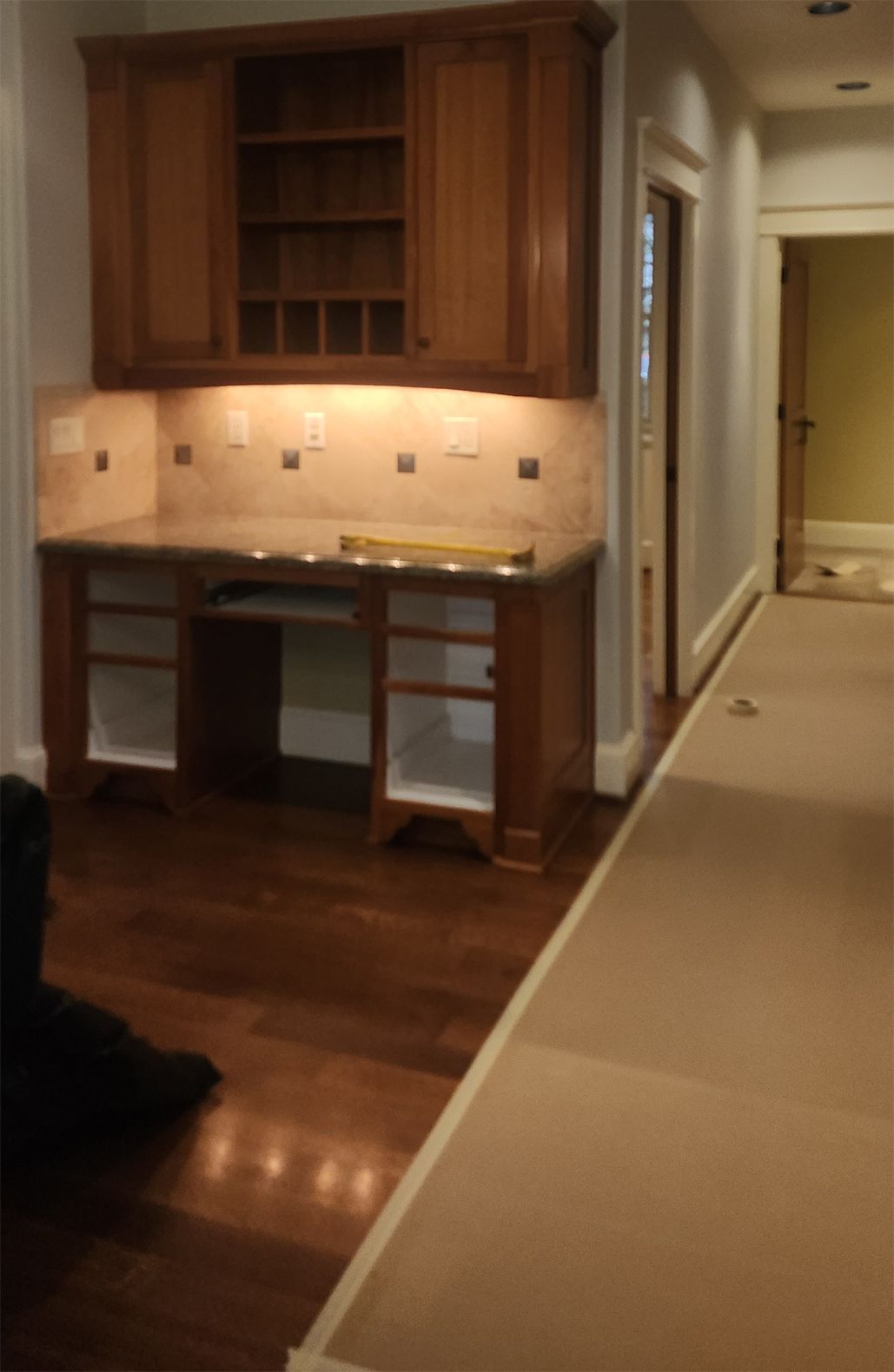 Built-in wooden desk and cabinet with overhead lighting, a wine rack, and a tiled backsplash in a room with hardwood floors.
