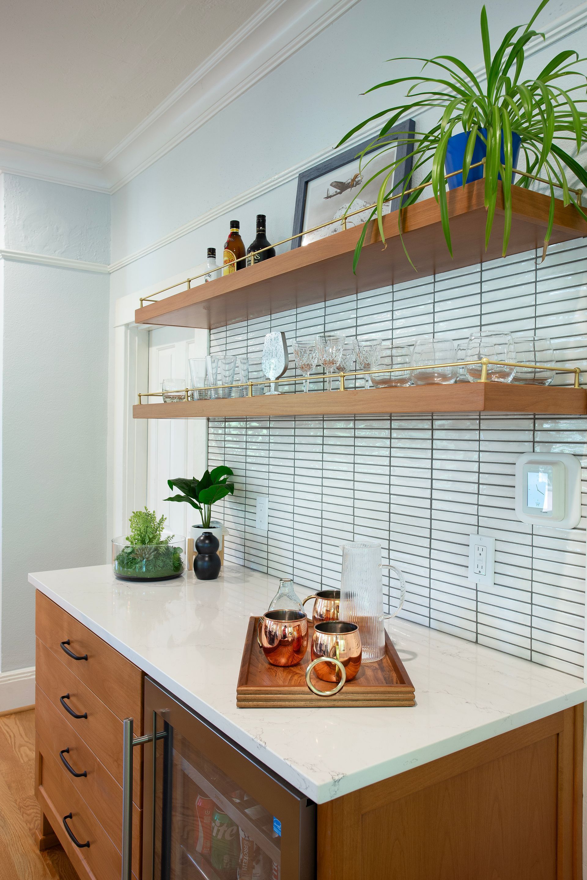 Wooden bar cabinet with marble top, shelves with glassware, and plants. Copper mugs on tray.