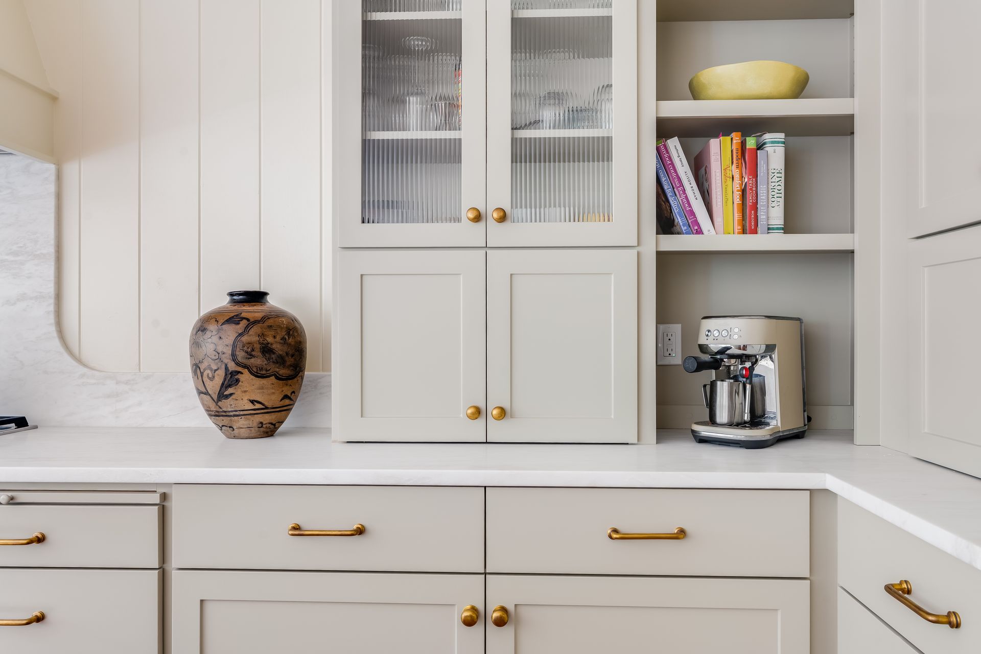 Kitchen cabinets with glass-fronted upper cabinet and built-in coffee machine, light gray and white with gold hardware.