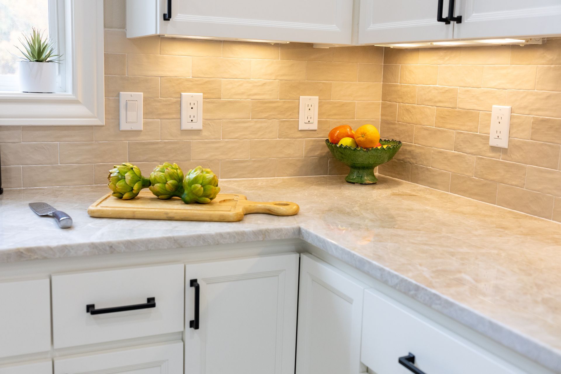 Kitchen countertop with artichokes on a cutting board, fruit bowl, white cabinets, and backsplash with overhead lighting.