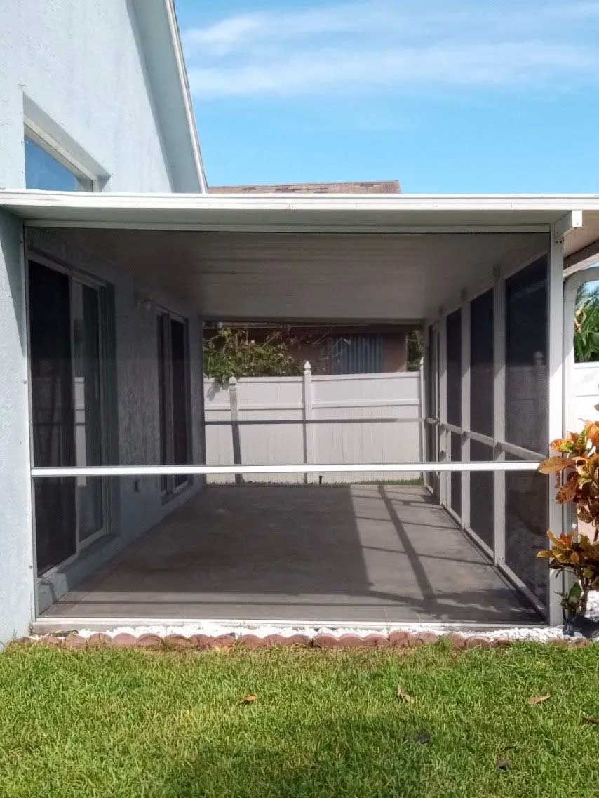 Screened-in patio with a white frame, attached to a light blue house, overlooking a grassy yard under a blue sky.
