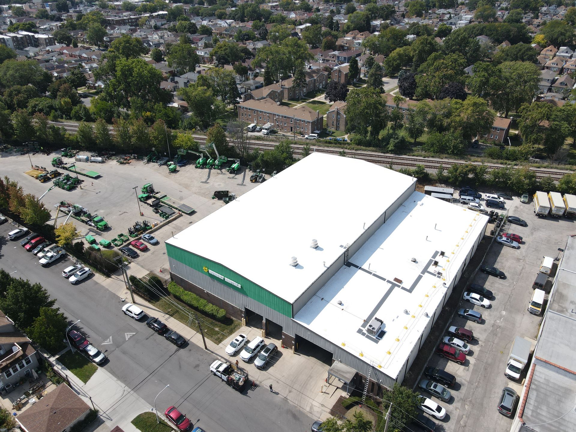 Aerial view of a large warehouse with a white roof and green trim; surrounded by parked vehicles and green equipment.