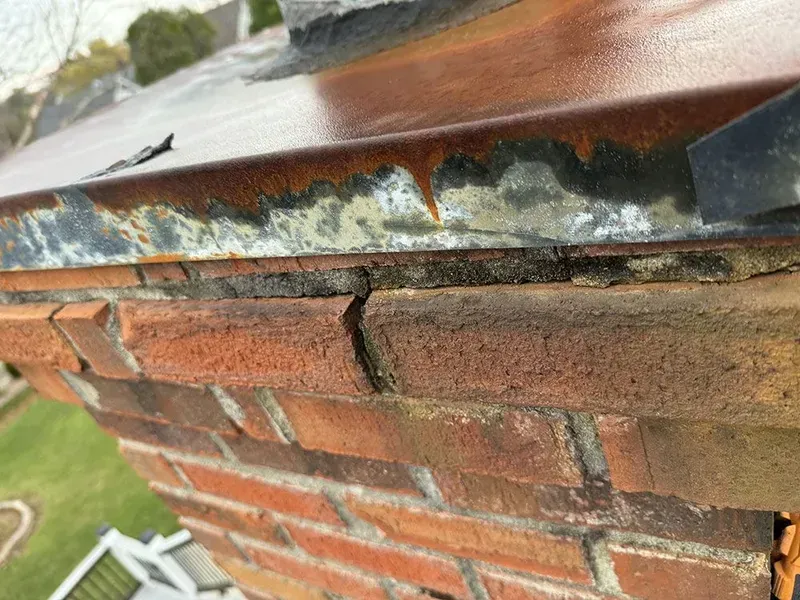 Close-up of a brick chimney showing rust on the metal flashing and cracks in the mortar.