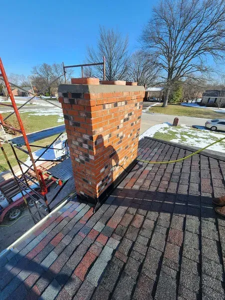 Brick chimney on a roof with visible asphalt shingles; a ladder is visible beside the chimney.
