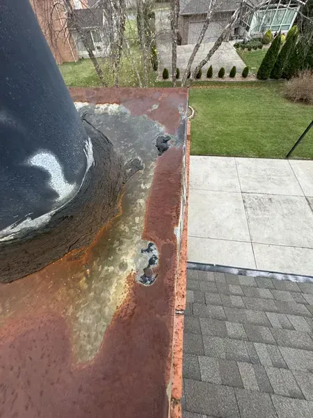 Close-up of a chimney top with rust and deterioration. A black flue pipe is attached, with a gray roof visible.