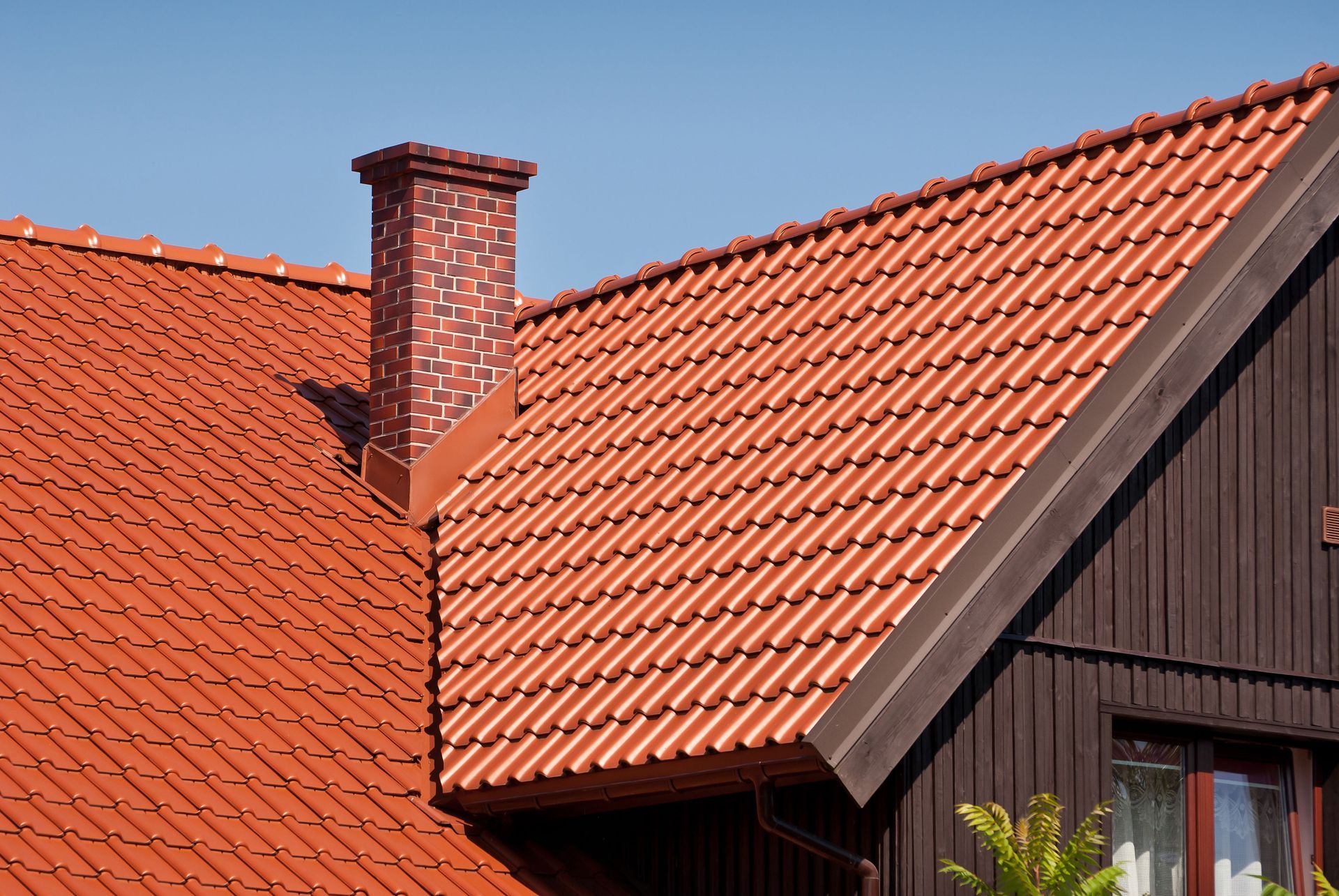 Red tile roof with brick chimney against a blue sky, partially showing brown siding.