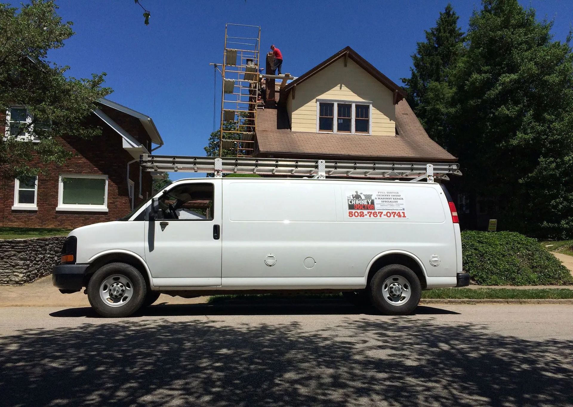 White work van parked in front of a house. Workers on roof and scaffolding, blue sky.