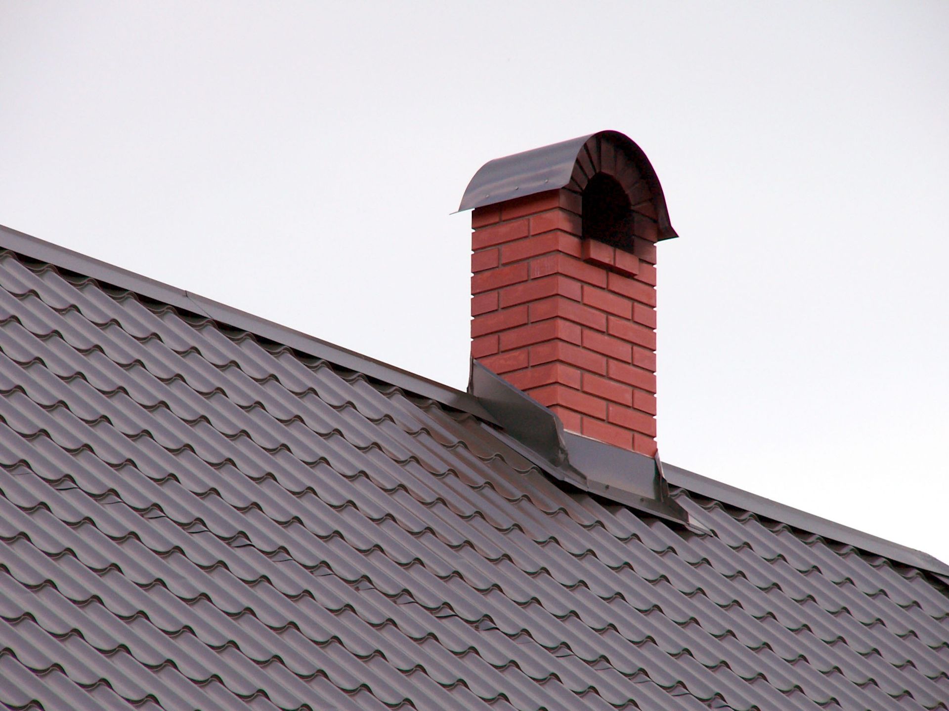 Red brick chimney on a gray metal roof, against a pale sky.