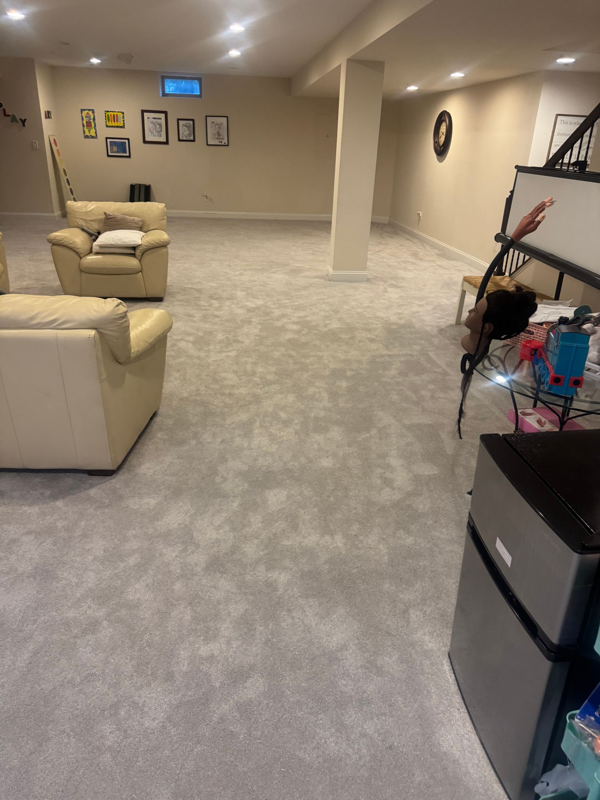 Light-colored basement room with neutral carpet, two cream armchairs, and a mini-fridge.
