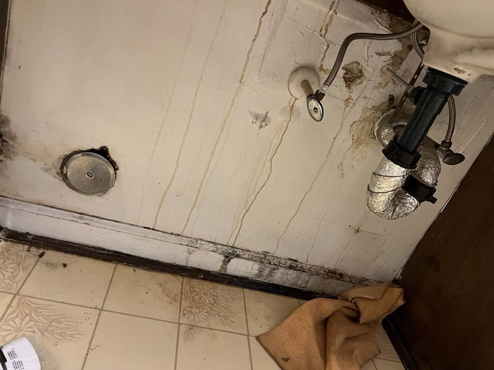 Underside of a dirty bathroom sink with visible water damage and plumbing. Brown and white tones.