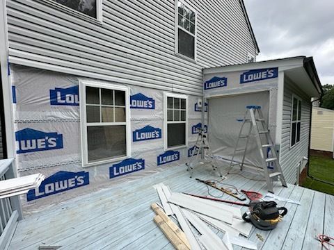 House exterior under renovation; siding removed, covered in Lowe's wrap. Deck with tools, ladders, and materials.