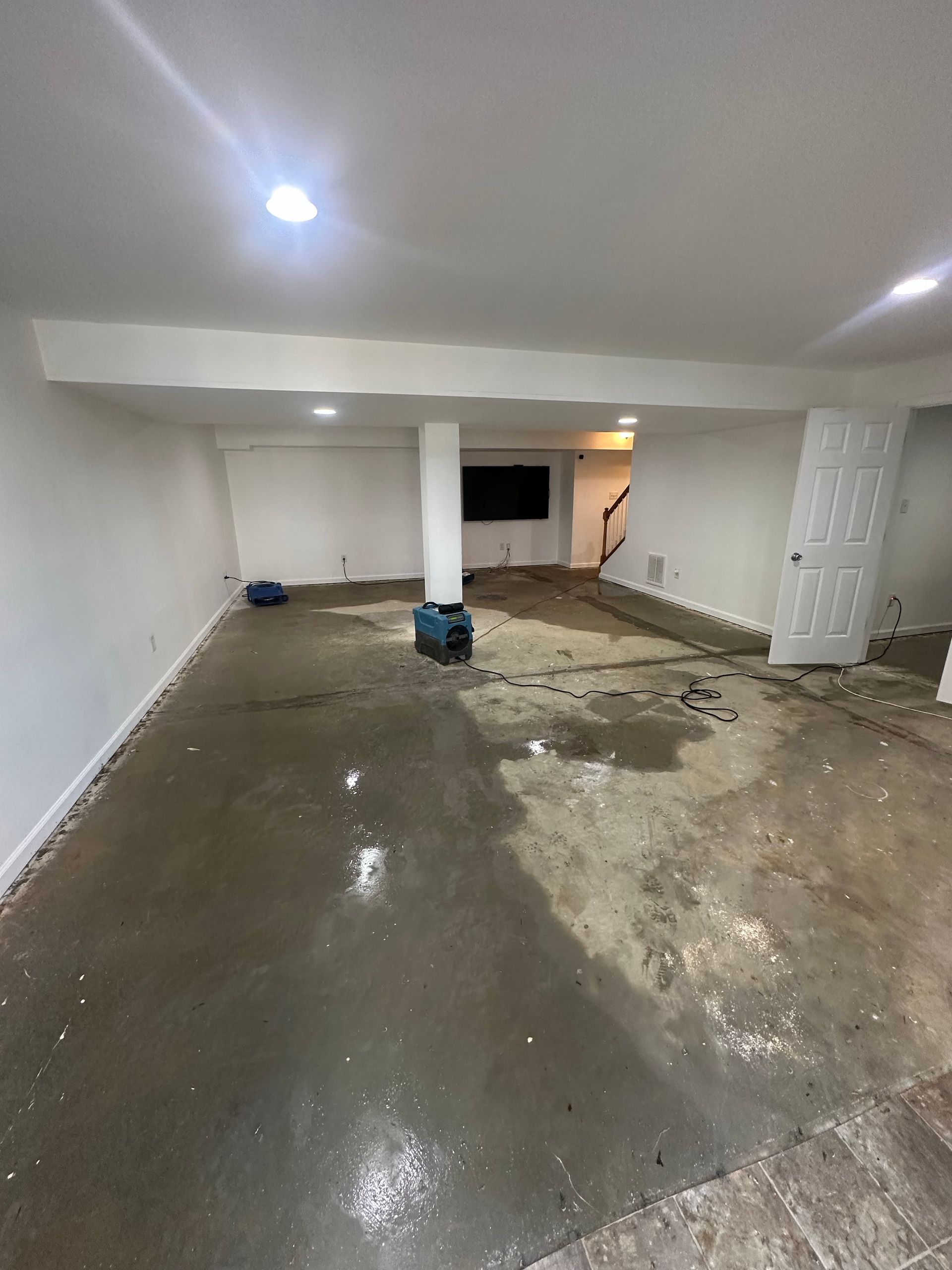 Basement with wet concrete floor, white walls, support beams, overhead lighting, and a door.